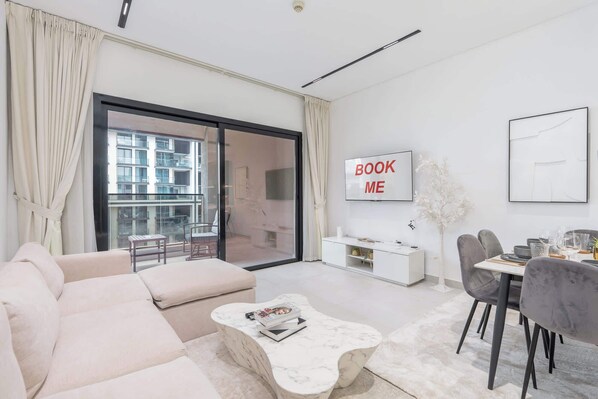 A modern living room with a sofa, a coffee table, a dining area with chairs, a TV, and a balcony with a view of buildings. - StayShort - Comfy JVC Retreat for 2 with Pool & Gym (JVC)