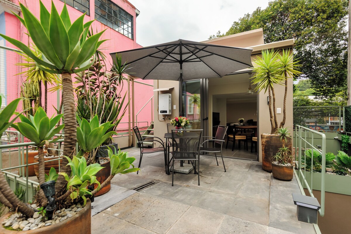 An outdoor patio area with a table and chairs under a large umbrella, surrounded by potted plants and a pink building in the background.