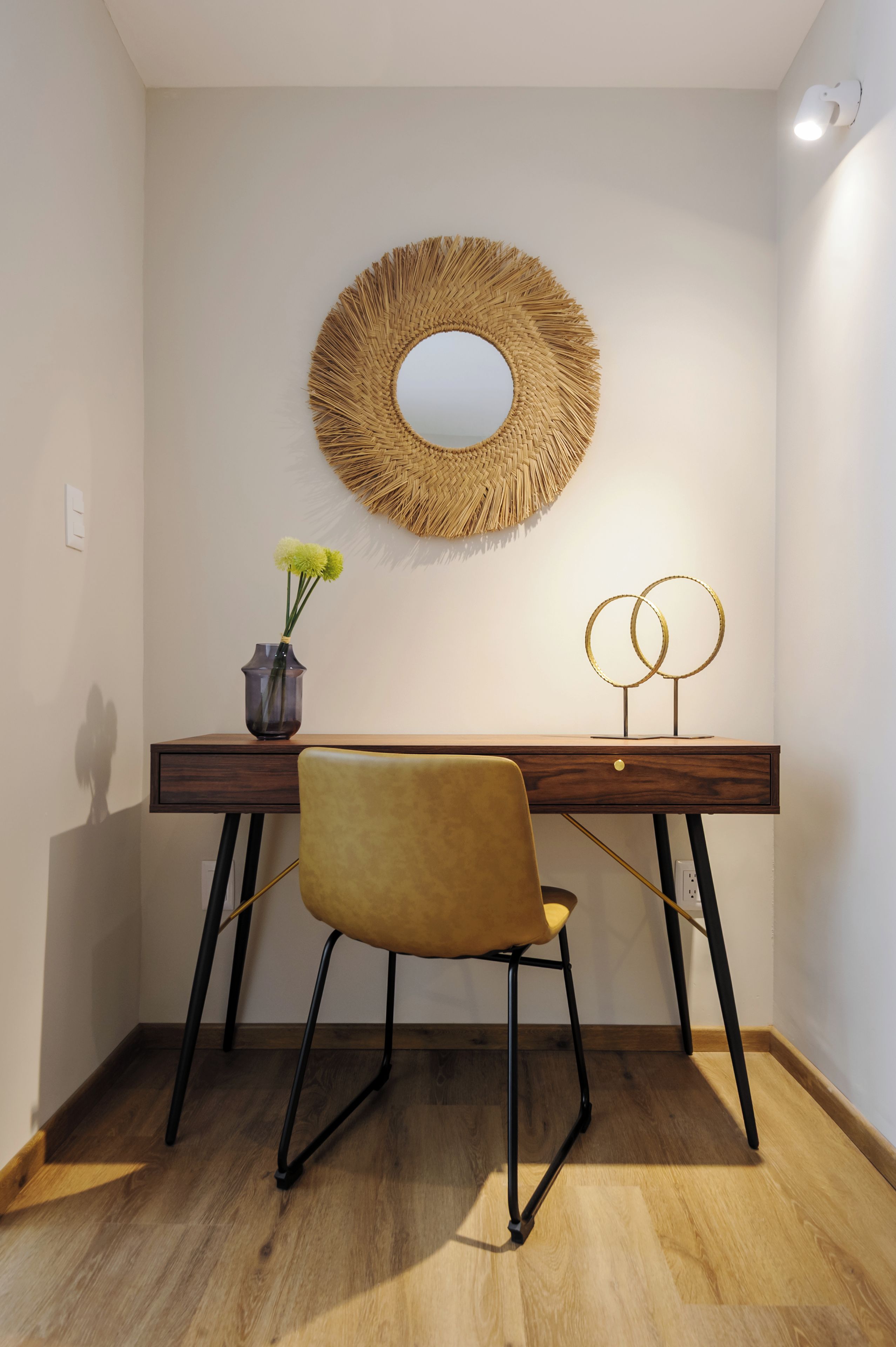 A desk with a woven mirror, a vase with flowers, and a gold lamp.