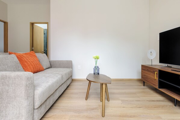 A modern living room with a grey sofa, a small wooden side table, a television on a wooden stand, and a door leading to another room. - Zaterra (Mexico City)