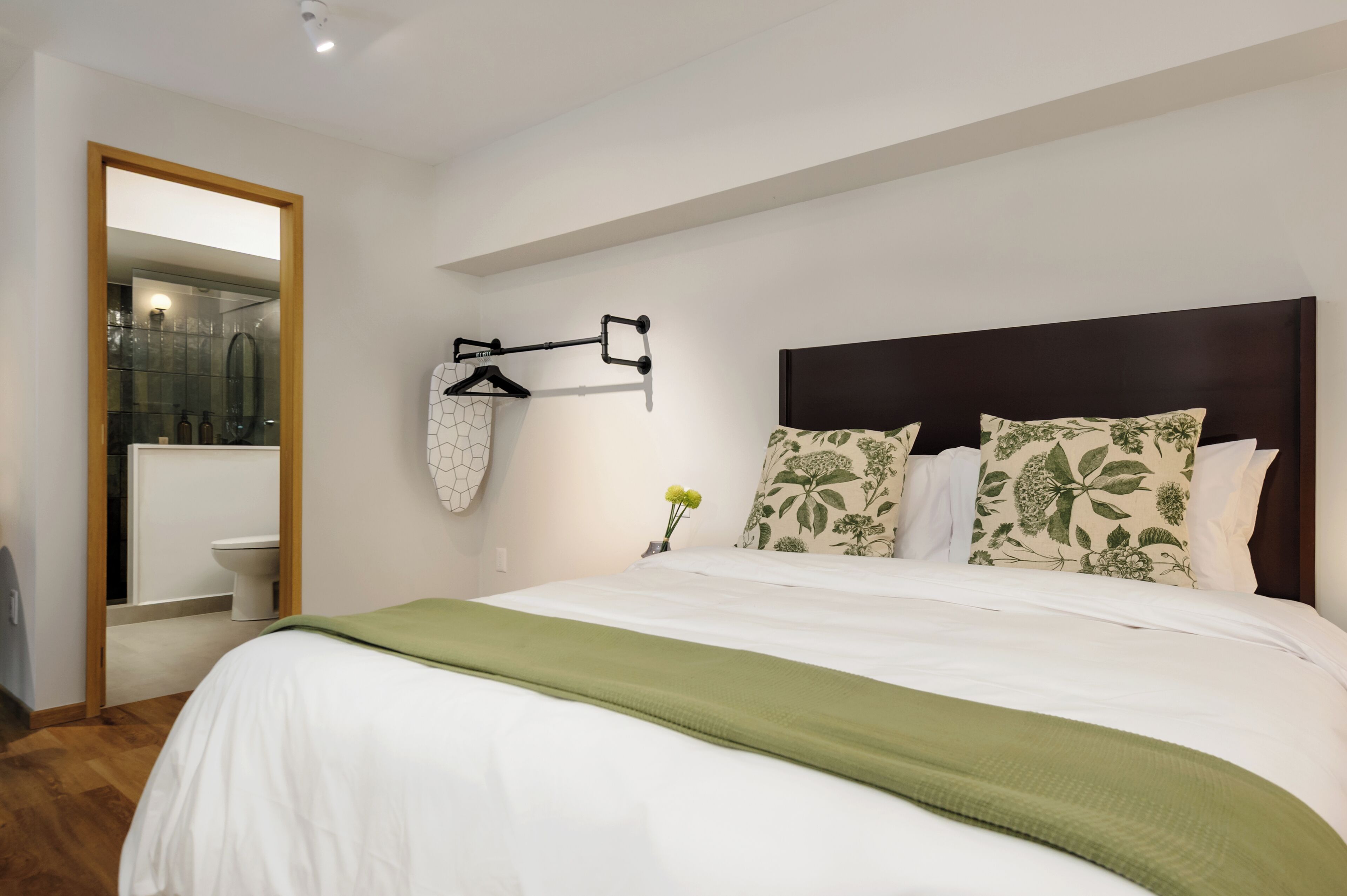 Een moderne hotelkamer met een groot bed, een nachtkastje met een bloevas, een wandrek voor handdoeken en uitzicht op de badkamer.