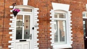 A two-story brick house with a white front door, two windows with shutters, and a small flower basket on the door. - Traditional Belfast House 10 Mins From City Centre (Belfast)