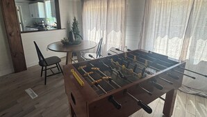 Ett rum med trägolv, ett fotbollsspel, ett matbord med stolar, en köksdel och ett fönster med gardiner.