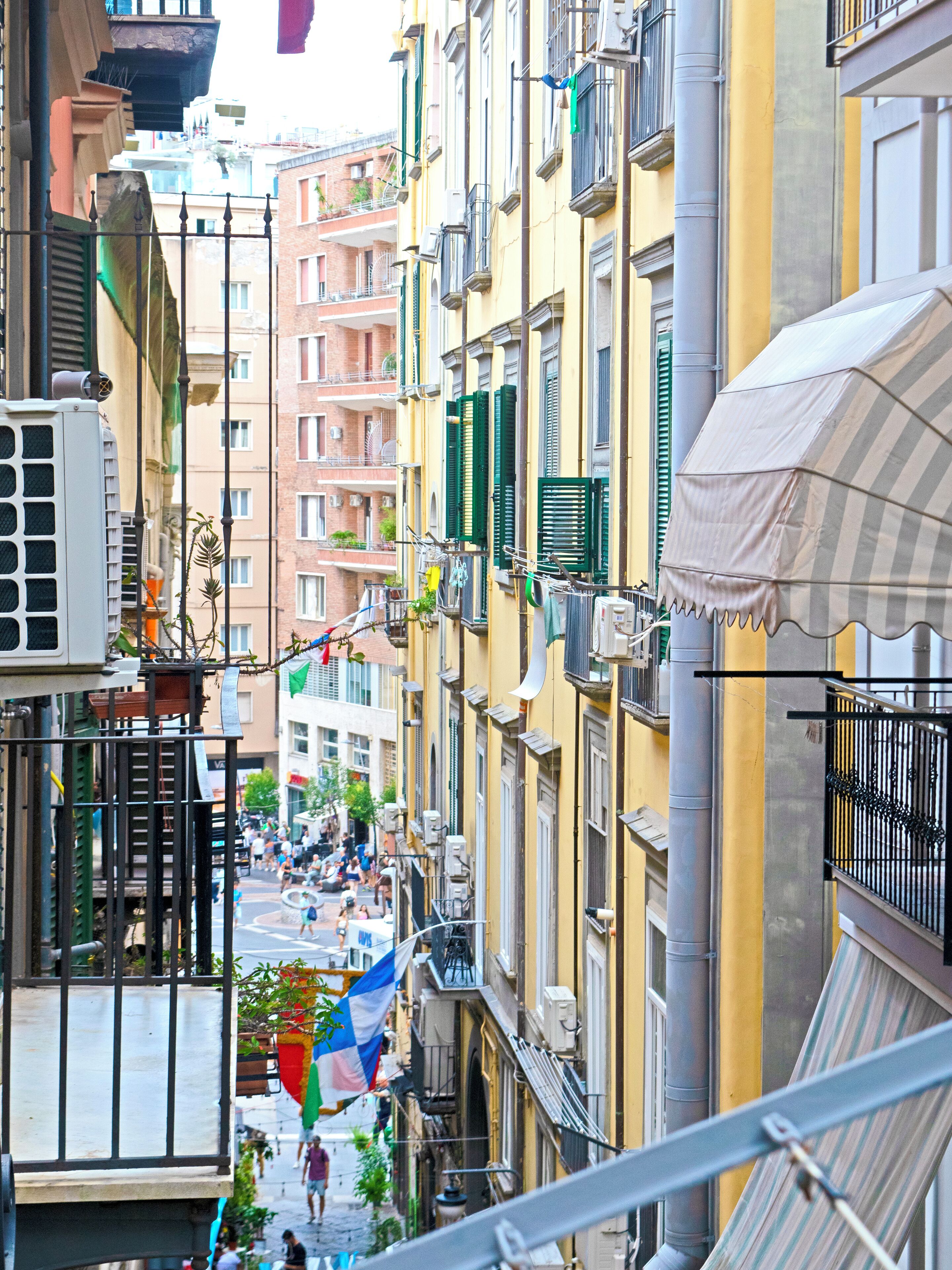 A narrow street with buildings on either side, balconies with laundry hanging, and a flag.