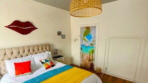 Quarto com cama, luminária pendente, arte na parede com lábios, porta com cena tropical e mesinhas de cabeceira.