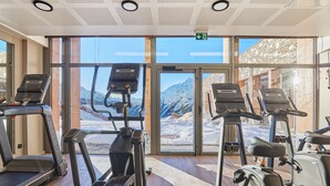 Ein Fitnessraum mit Laufbändern, Crosstrainern und einem großen Fenster mit Blick auf die Berge.