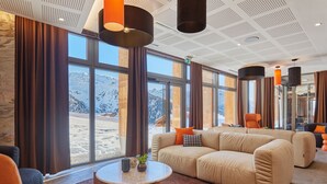 Ein modernes Hotelzimmer mit großem Fenster und Blick auf die Berge, einer beigefarbenen Couch, einem runden Couchtisch und einer Sitzecke mit einem Sessel.