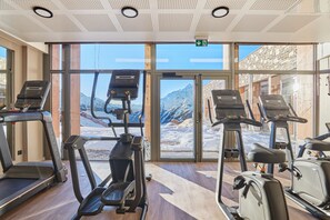 Een fitnessruimte met loopbanden, een crosstrainer en een hometrainer, grote ramen met uitzicht op de bergen.