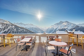 A terrace with tables and chairs, overlooking snow-covered mountains under a clear sky.