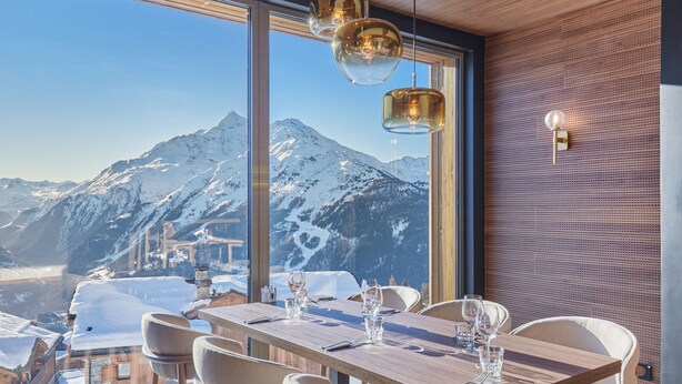 A dining area with a table set for four, large windows offering a mountain view, and pendant lighting. - Suite Familiale | À 100m des Pistes | Piscine (Montvalezan)