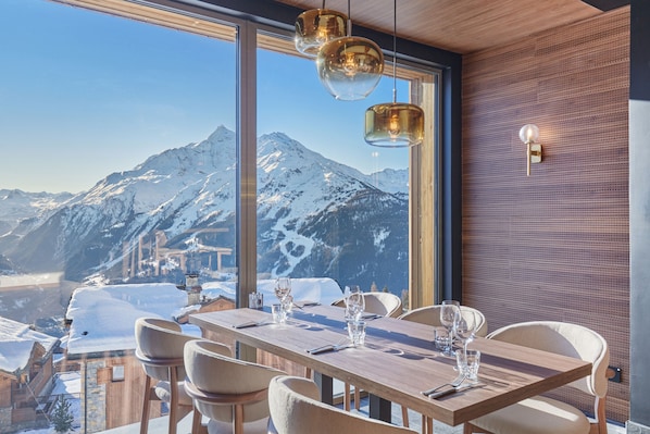 A dining area with a table set for four, large windows offering a mountain view, and pendant lighting. - Suite Familiale | À 100m des Pistes | Piscine (Montvalezan)