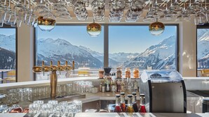 A bar with multiple glasses and bottles, a bar tap, and a view of snow-capped mountains through large windows. - Suite Familiale | À 100m des Pistes | Piscine (Montvalezan)