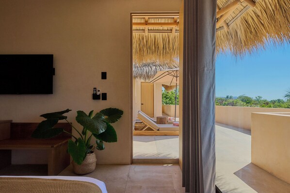 A room with a bed, a plant, a TV, and a view of a balcony with a thatched roof. - Casa Cambria (Santa Maria Colotepec)