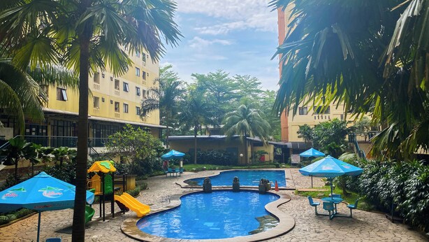 A swimming pool area with palm trees, a children's play area, and blue umbrellas. - Homey 2Br With Modern Design Kebagusan City Apartment (Jakarta)