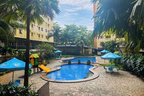 A swimming pool area with palm trees, a children's play area, and blue umbrellas. - Homey 2Br With Modern Design Kebagusan City Apartment (Jakarta)