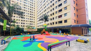 A playground with colorful play equipment and a soft play surface, surrounded by a high-rise residential building. - Homey 2Br With Modern Design Kebagusan City Apartment (Jakarta)