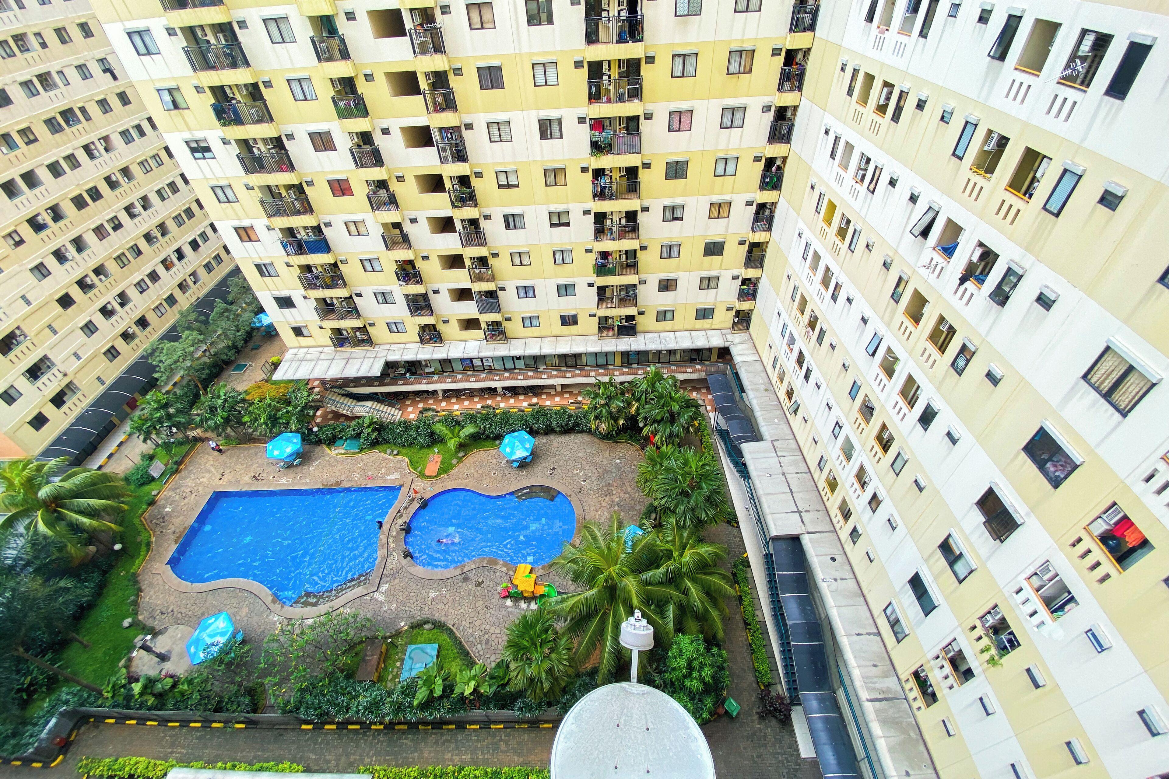 A high-rise residential building with multiple balconies, a central courtyard with two swimming pools, and surrounding greenery.