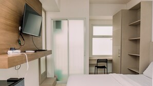 A hotel room with a bed, a TV mounted on a wooden panel, a desk with a chair, and a wardrobe. - Fully Furnished And Simply Studio At Serpong Garden Apartment (Cisauk)