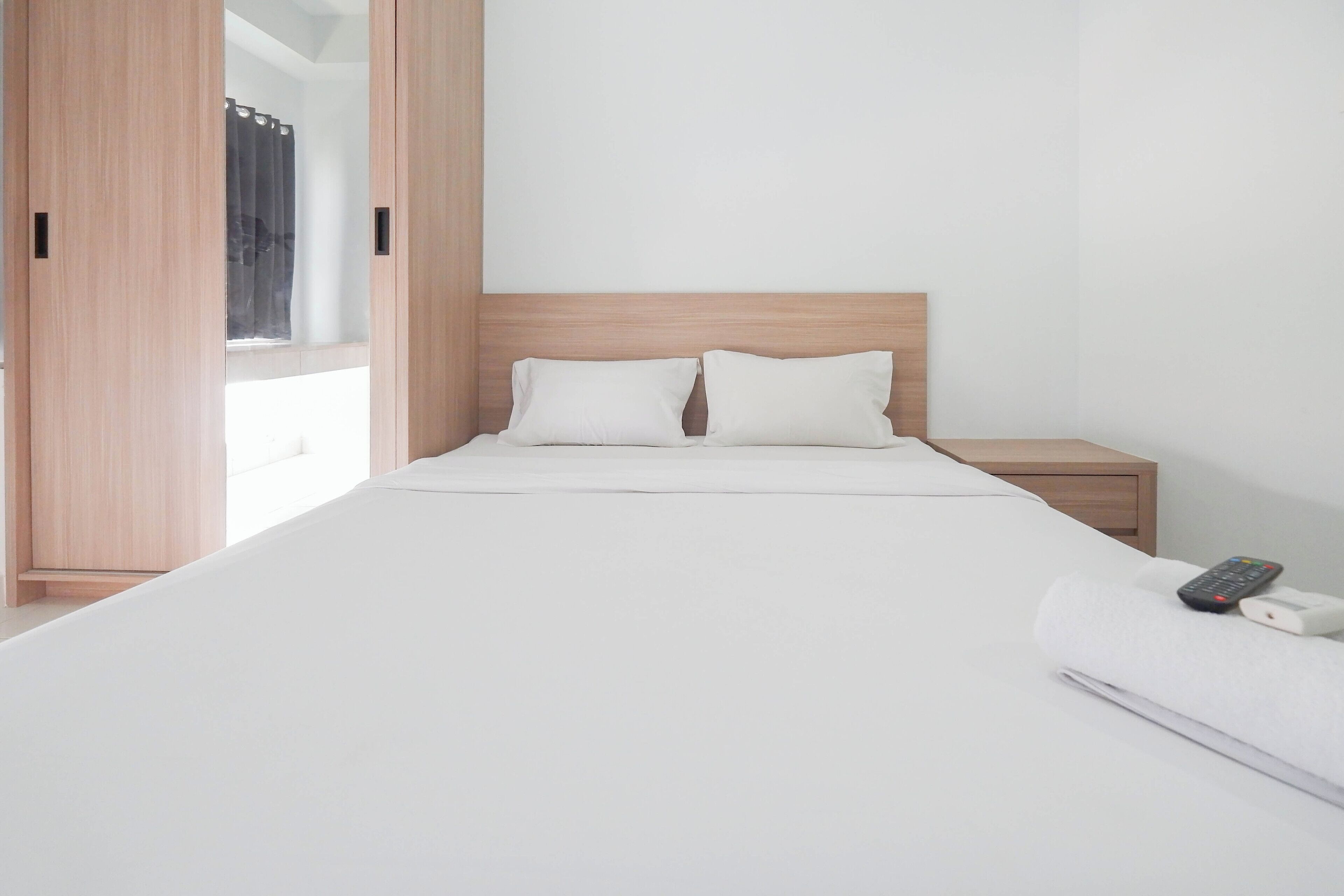 Minimalistinen hotellihuone, jossa on suuri sänky, kaksi tyynyä, puinen pääty, yöpöytä, jossa on kaukosäädin, ja osittain vedettynä oleva verho.