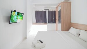 A hotel room with a bed, a TV mounted on the wall, a window with curtains, and a wooden wardrobe. - Studio Patraland Urbano Apartment (Bekasi)