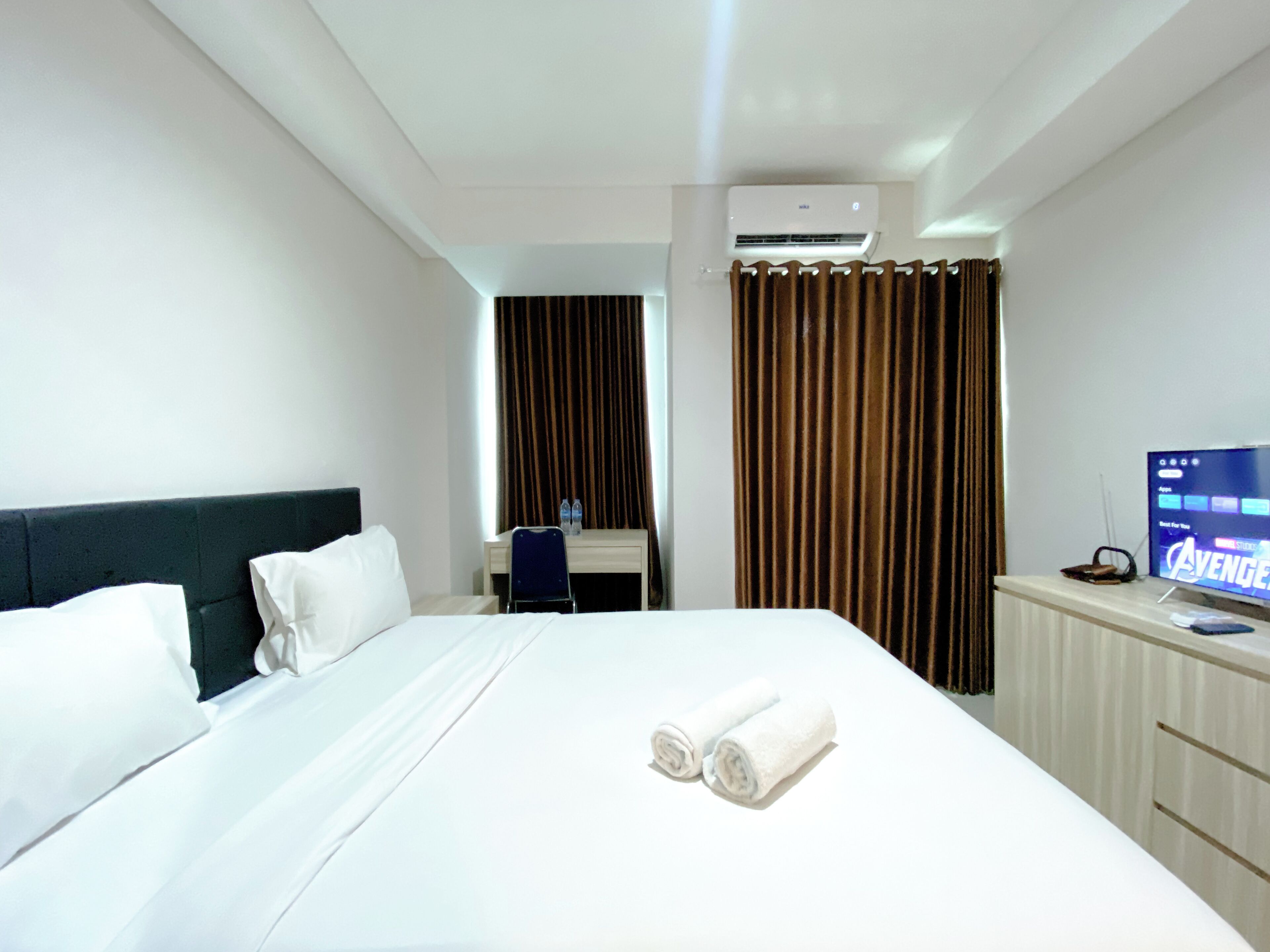A hotel room with a bed, a desk, a chair, a TV, and an air conditioning unit.