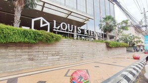 The exterior of Louis Kienne Pemuda hotel with a modern facade, a landscaped area with palm trees, and a clear sky. - Strategic And Homey Studio At Marquis De Lafayette Apartment (Semarang)