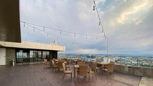 An outdoor terrace with wooden tables and chairs, string lights, and a city view. - Strategic And Homey Studio At Marquis De Lafayette Apartment (Semarang)