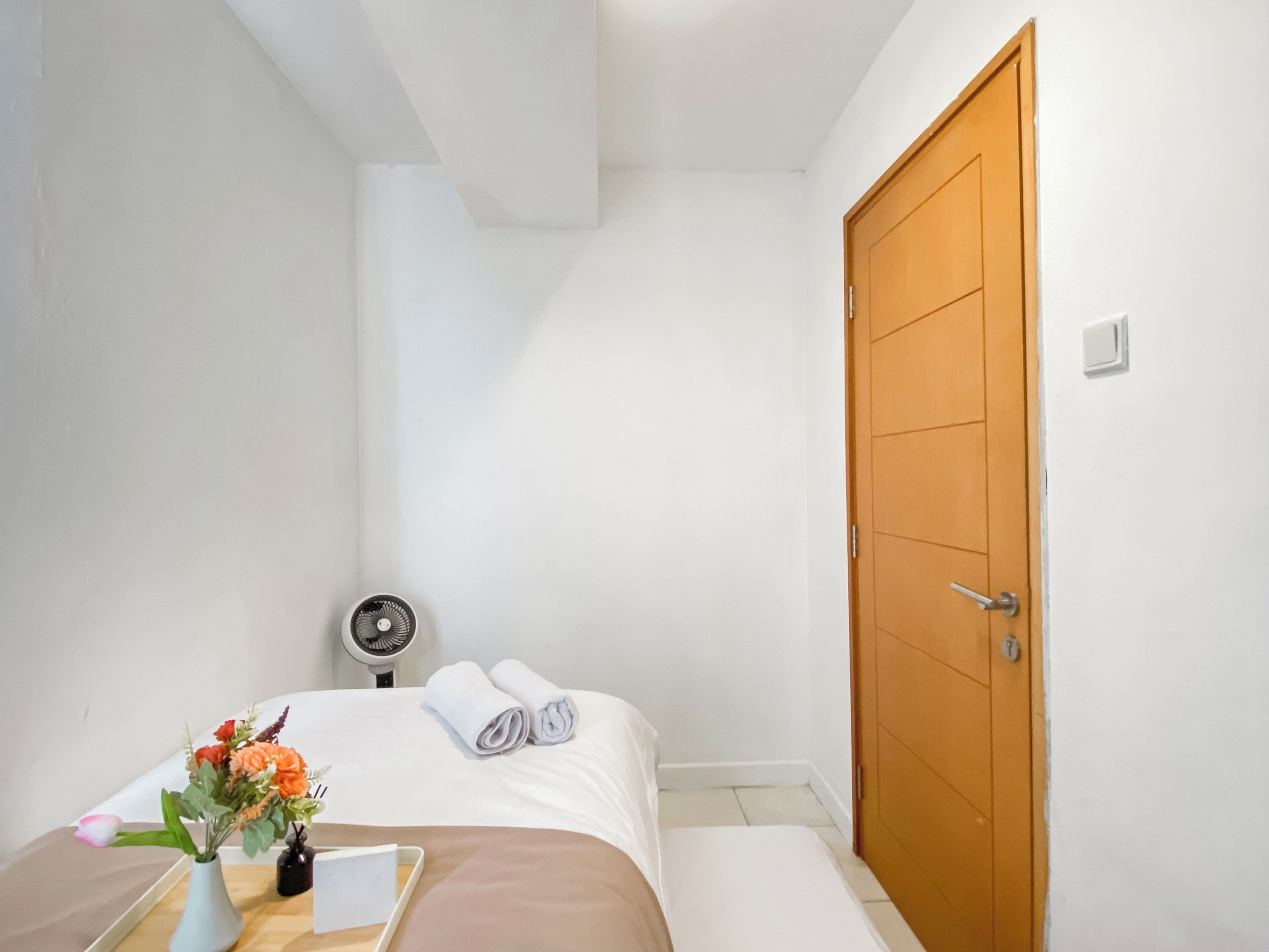 Une petite pièce propre, avec un lit, une table de chevet ornée d’un vase de fleurs, un ventilateur et une porte en bois.