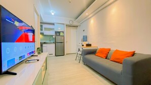 A modern living room with a flat-screen TV, grey sofa with orange cushions, and a kitchen in the background. - Warm And Cozy Stay 2Br Pakubuwono Terrace Apartment (Jakarta)