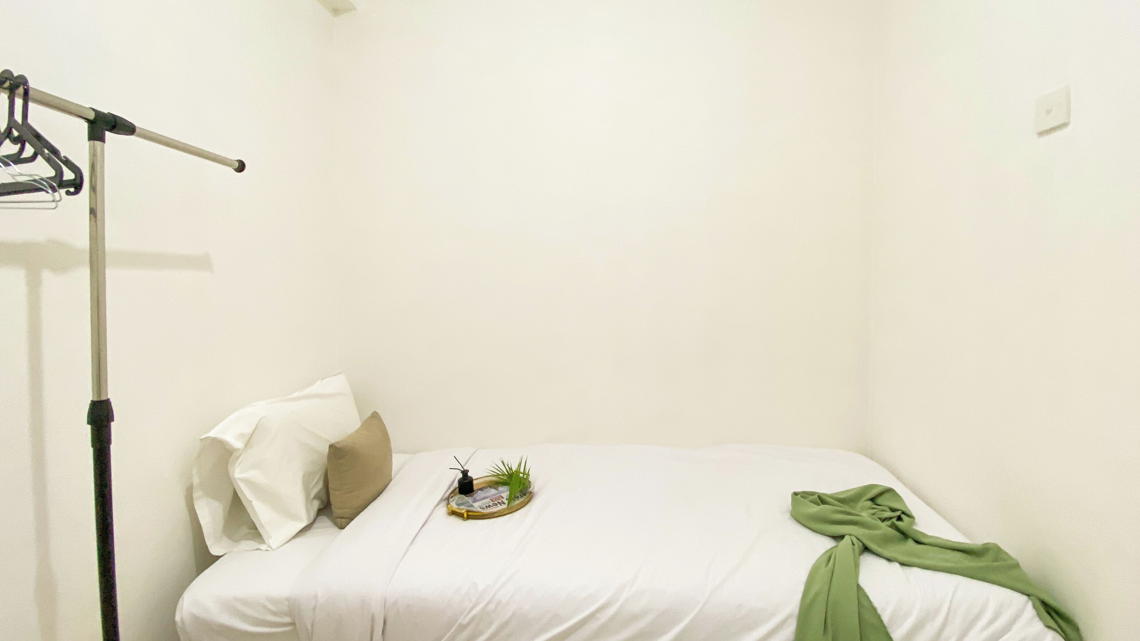 Un petit lit bien fait, avec une literie blanche et un tissu vert drapé dessus. On aperçoit une petite plante sur le lit et un objet noir, qui pourrait être une télécommande, posé sur un plateau. Un cintre noir est fixé au mur, à gauche.