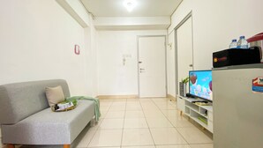 A living room with a grey sofa, a flat-screen TV, a refrigerator, and a cabinet. - Homey And Modern 2Br At Green Bay Pluit Apartment (Jakarta)