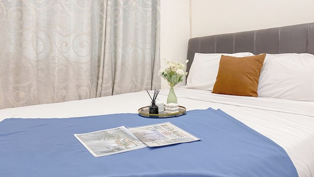 A neatly made bed with a blue bedspread, white pillows, and a brown accent pillow. A vase with white flowers and a tray with a newspaper and a pen holder are on the bed. - Good Place And Comfortable 2Br At Green Pramuka City Apartment (Jakarta)