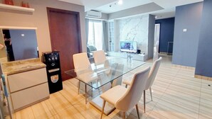 Una sala de estar moderna con una mesa de comedor de vidrio, sillas blancas, un televisor y una zona de cocina con refrigerador y máquina expendedora.