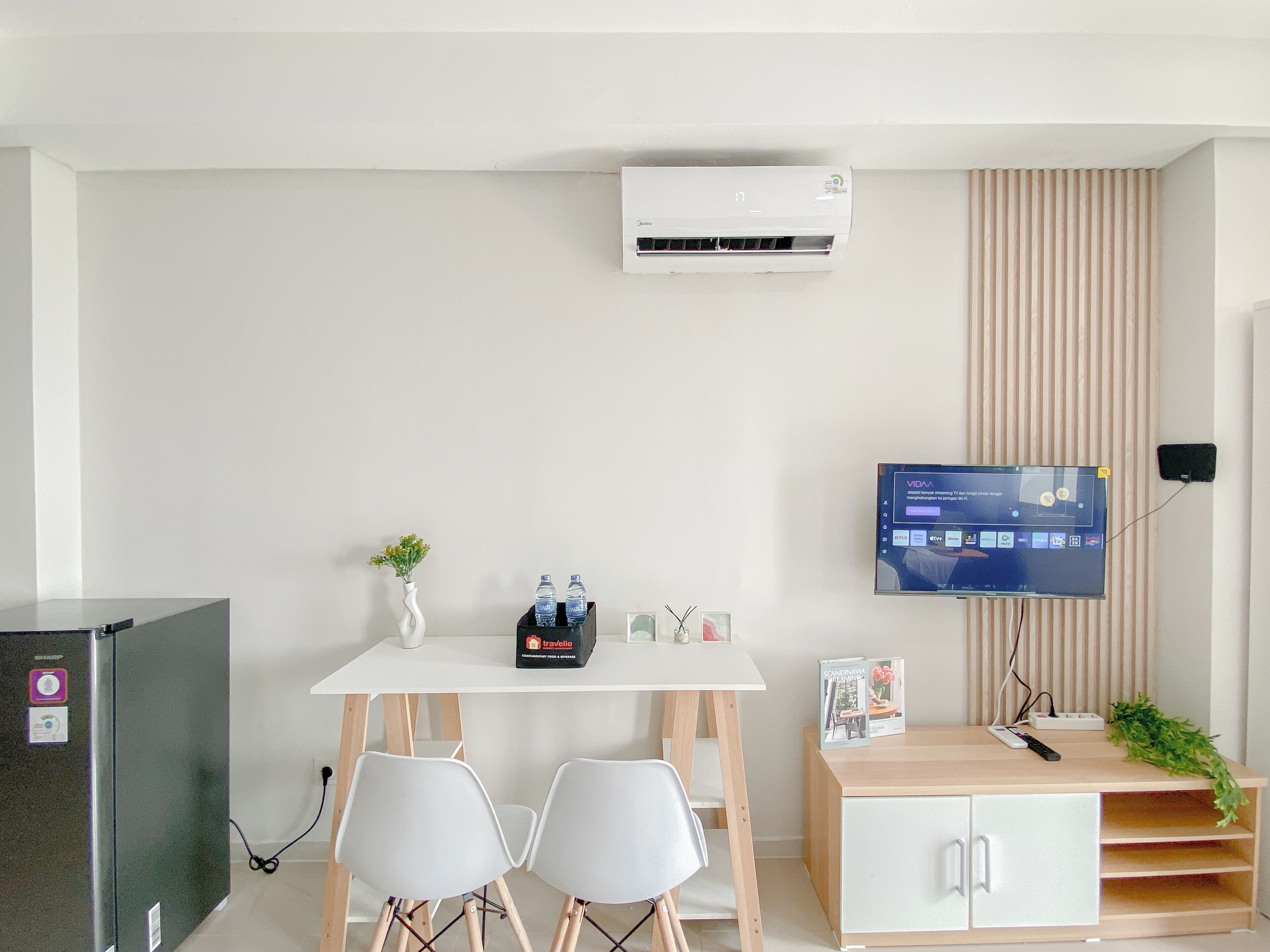 Ein modernes, minimalistisches Zimmer mit weißem Schreibtisch, zwei Stühlen, einem kleinen Tisch mit Vase und Wasserflaschen, einem an der Wand montierten Fernseher und einem kleinen Schrank mit Fernbedienung.
