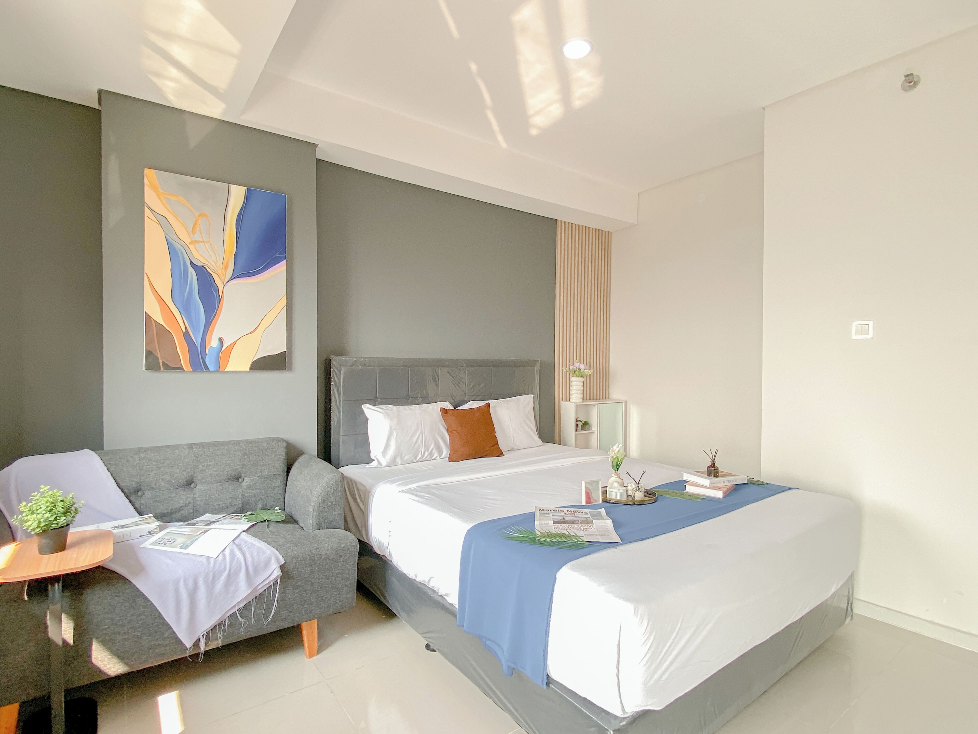 A modern hotel room with a grey sofa, a large bed with white bedding, a decorative abstract painting, and a small table with a plant and a newspaper.