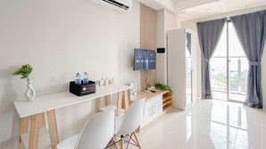 A modern hotel room with a white desk, two white chairs, an air conditioning unit, a flat-screen TV, a small plant, and a window with curtains. - Cozy Studio Daan Mogot City Apartment (Jakarta)