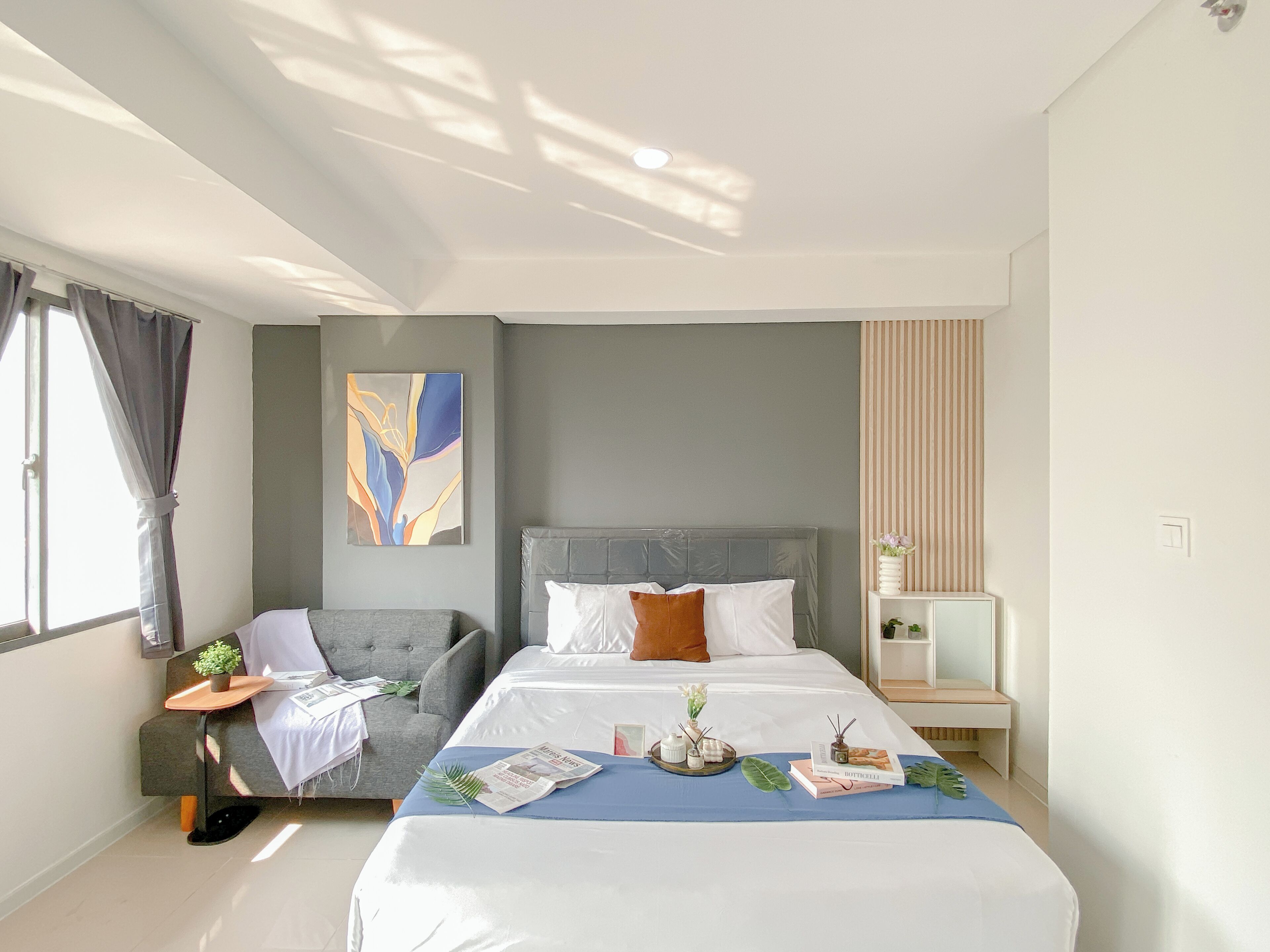 A modern hotel room with a large bed, a grey armchair, a small side table, and a decorative wall piece.