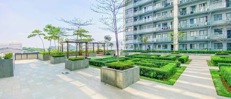 A modern residential building with multiple balconies, a landscaped courtyard with neatly trimmed hedges, and a series of planters with trees.