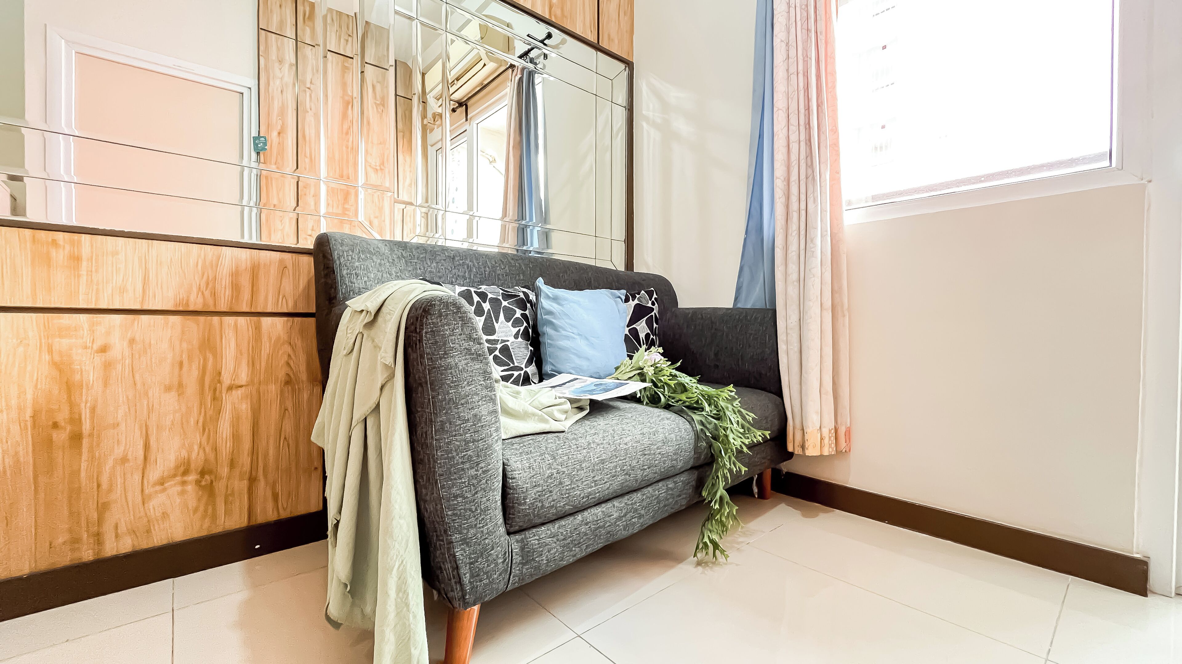 A modern living room with a grey sofa, wooden cabinets, a large mirror, and a window with curtains.