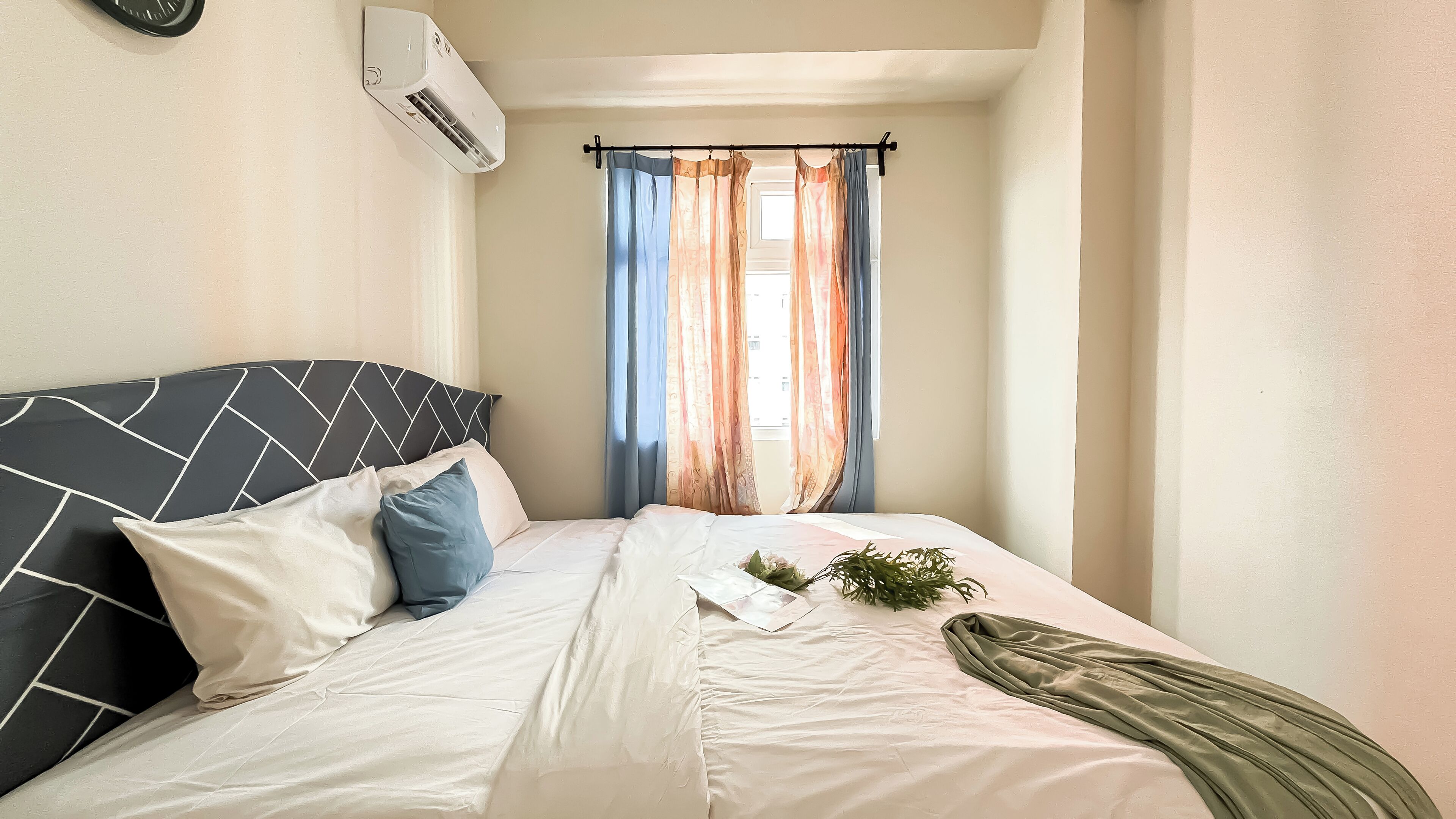 Ein ordentlich bezogenes Bett mit grau und weiß gestreifter Kopfteil, weißer Bettwäsche und einer grünen Tagesdecke. Ein Fenster mit orange-blauen Vorhängen, und eine Klimaanlage ist an der Wand montiert.