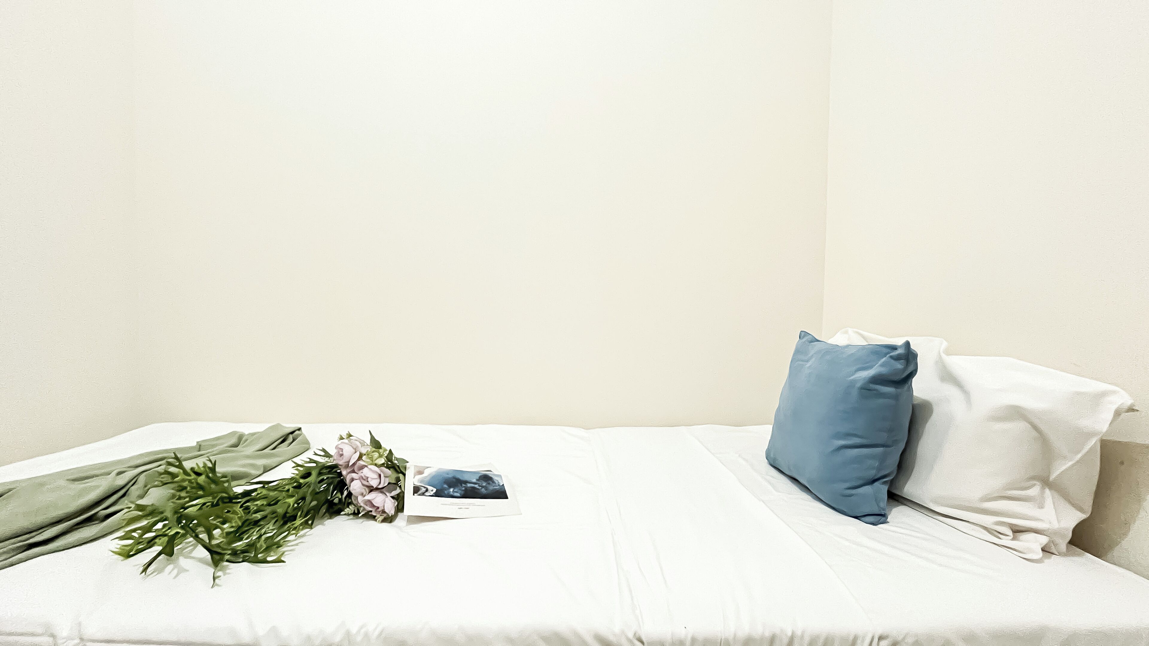 A single bed with a white sheet, a blue pillow, and a bouquet of flowers.