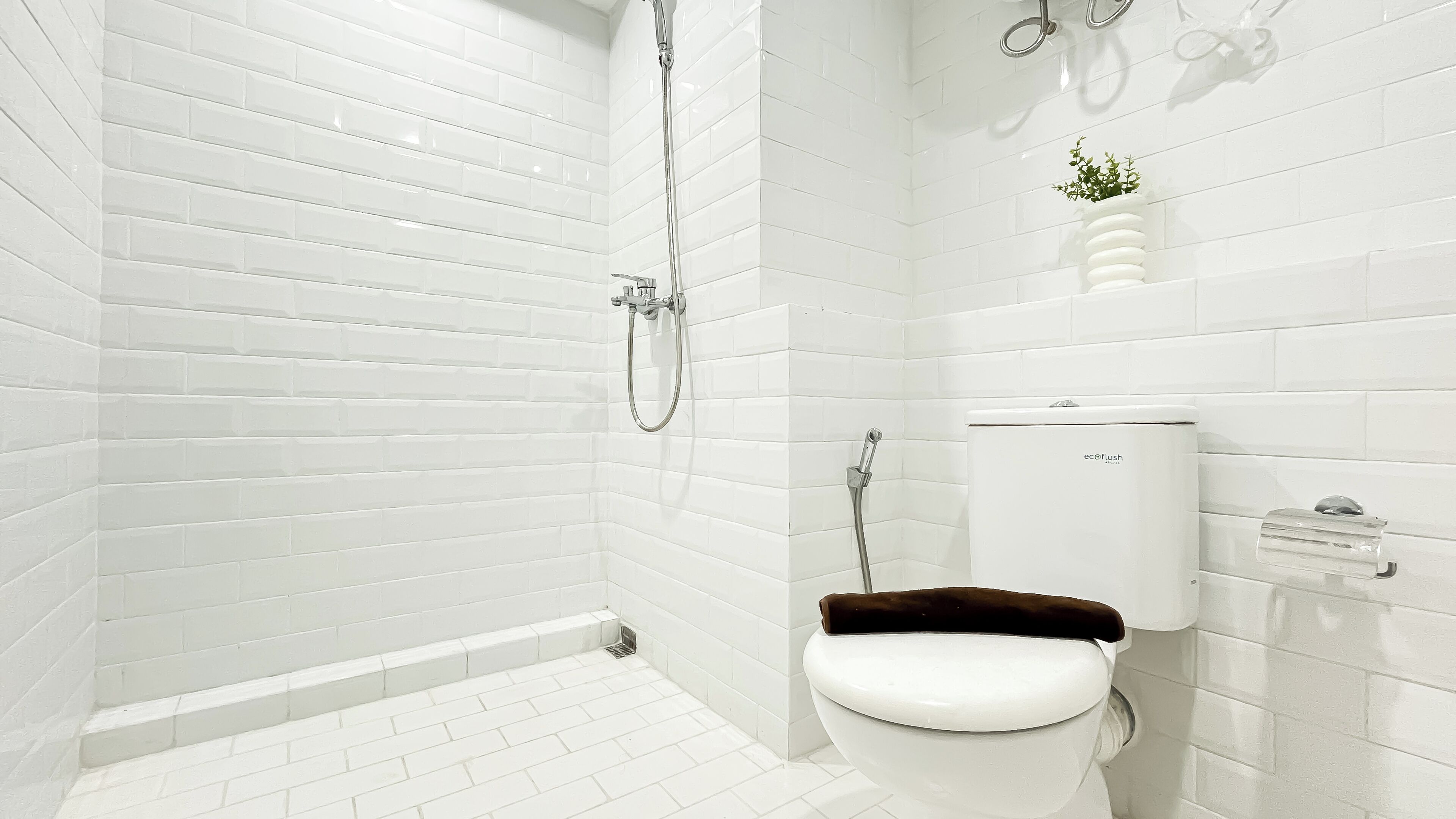 Un baño moderno con azulejos blancos tipo metro, un inodoro blanco, una ducha con cabezal de ducha manual y una pequeña planta en maceta en la pared.