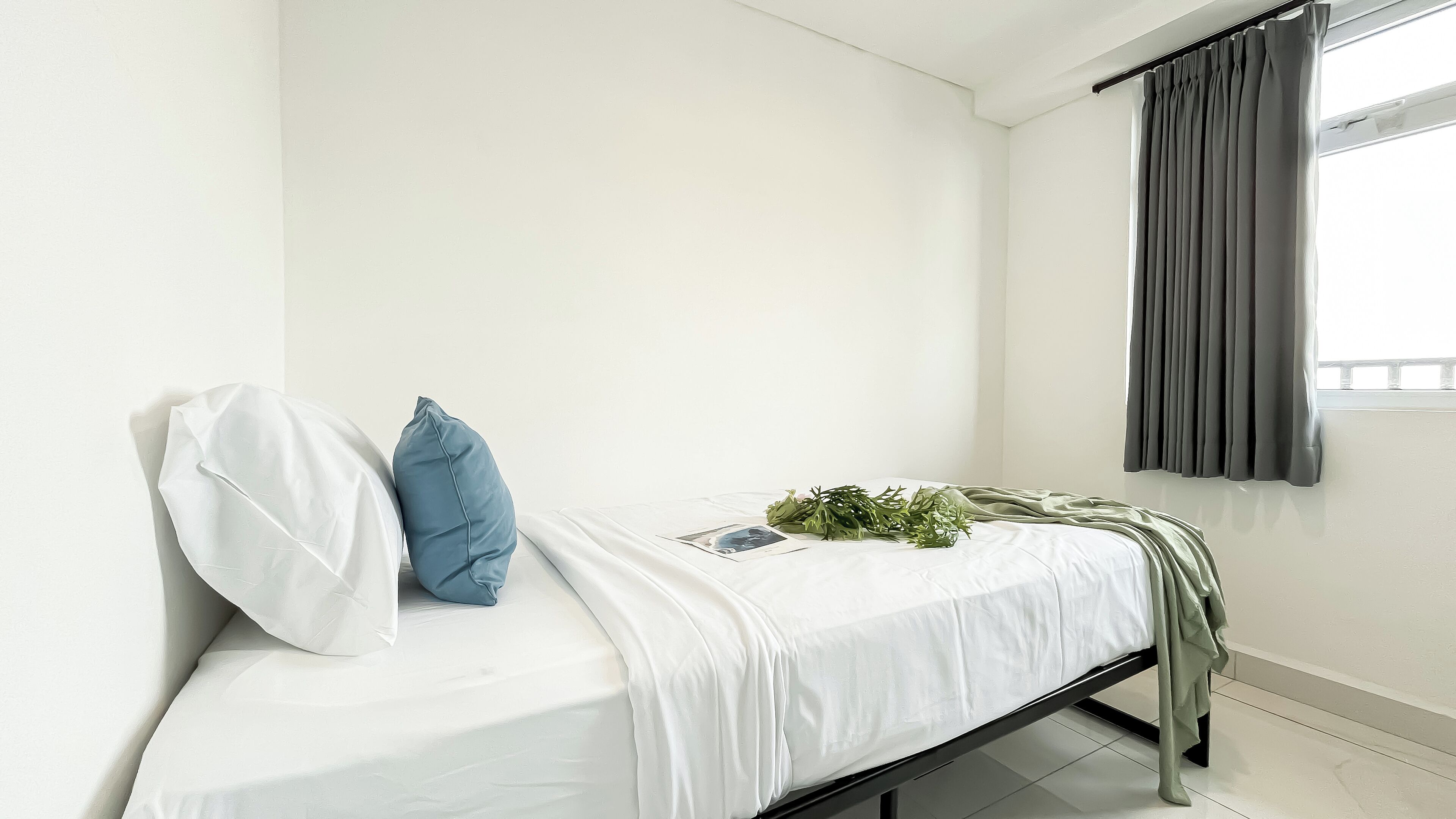 A single bed with white bedding, a blue pillow, and a green plant.