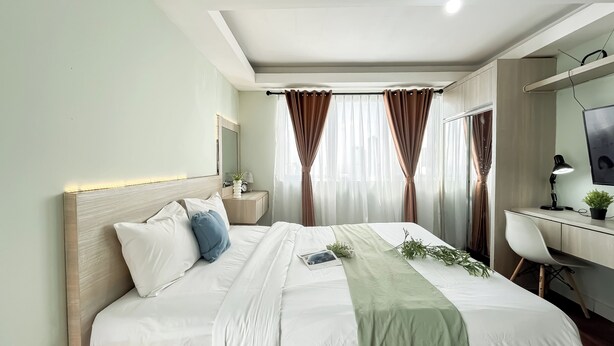 Ein modernes Schlafzimmer mit einem ordentlich bezogenen Bett, einem Schreibtisch mit Lampe und einem großen Fenster mit Vorhängen.