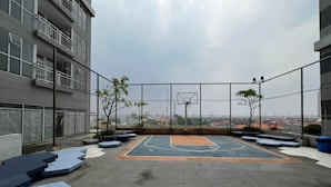 An outdoor basketball court with a net, surrounded by seating areas and trees, set against a cityscape backdrop. - Good Deal Stunning Studio (No Kitchen) At Taman Melati Surabaya Apartment (Surabaya)