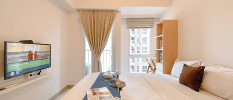 A hotel room with a bed, a television mounted on the wall, a window with curtains, a bookshelf, and a view of the outside buildings.