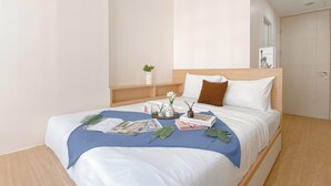 A neatly made bed with a blue bedspread, white pillows, and a brown accent pillow. There are decorative items on the bed including a vase with flowers, a small plant, and a few books. - Enjoy Living Studio At Tokyo Riverside Pik 2 Apartment (Teluknaga)
