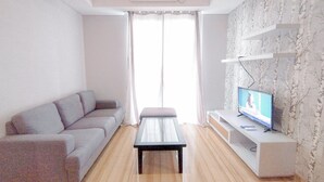 A modern living room with a grey sofa, a coffee table, and a TV on a white stand. - 2Br Minimalist And Simply At Grand Sungkono Lagoon Apartment (Surabaya)