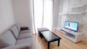 A modern living room with a grey sofa, a glass coffee table, and a TV on a white entertainment unit. - 2Br Minimalist And Simply At Grand Sungkono Lagoon Apartment (Surabaya)