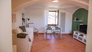 A cozy living room with a white sofa, a dining table with chairs, a TV on a white cabinet, and a window with curtains. - Cozy House with Lake View • Private Terrace & Self Check-in 24h (Viverone)